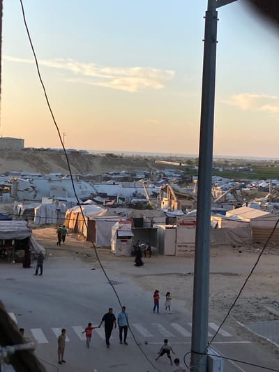 This image shows a view from above, possibly from a building, overlooking a refugee camp or densely populated area in Khan Yunis, Palestinian Territories. Numerous shelters and structures with white and light-colored roofs are visible. In the lower right corner, people are walking on a street or road with white crosswalk markings. Some individuals appear to be children. A metal pole is present in the foreground, obscuring part of the view. Power lines run across the scene. The sky is visible with orange and pale blue hues, suggesting either sunrise or sunset.