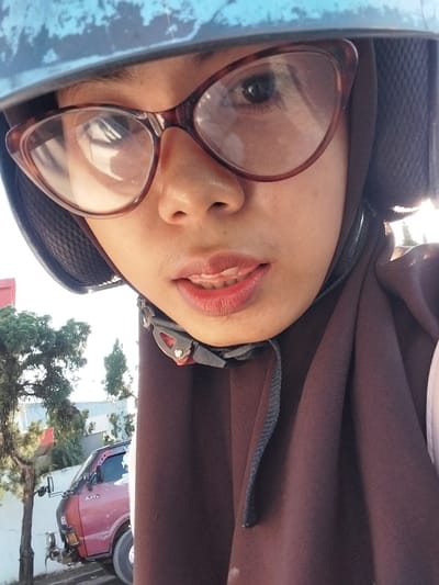 This is a close-up, low-angle selfie taken by a young woman wearing glasses and a dark brown hijab. She is inside a vehicle, possibly a car, as indicated by the blue interior in the foreground. The woman has her mouth slightly open, revealing her teeth and red lipstick. The image is taken outdoors during the day, with trees and a pink vehicle visible in the background. The pink vehicle appears to be a three-wheeled auto-rickshaw, common in Indonesia. The sky is bright, suggesting good weather. The overall mood of the image is casual and relaxed.