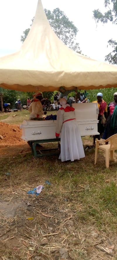 The image shows a funeral scene in Burundu, Kenya (latitude: 0.465, longitude: 34.74). There are people gathered under a large white tent. A white coffin is present on a wooden stand. A woman wearing a white dress and a red sash is standing beside the coffin. Other people are standing and sitting around the coffin, some wearing traditional clothing. There is a small pile of dirt in the foreground, and the rest of the ground is covered in grass and branches. A yellow chair sits beside the coffin. There is a white piece of fabric lying on the ground in the lower left of the image.