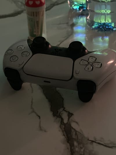 In Gwarinpa, Nigeria, a white gaming controller is placed on a marble-patterned table. The controller is a PlayStation 5 DualSense controller, and its buttons and joysticks are visible. In the background, partially out of focus, are two clear plastic bottles filled with a blue and green liquid, possibly beverages. To the left of the controller, a small cylindrical container with a red top decorated with white hearts and filled with what appear to be toothpicks or cotton swabs is also present. The lighting in the scene is dim, with reflections from the bottles casting colored light onto the table.