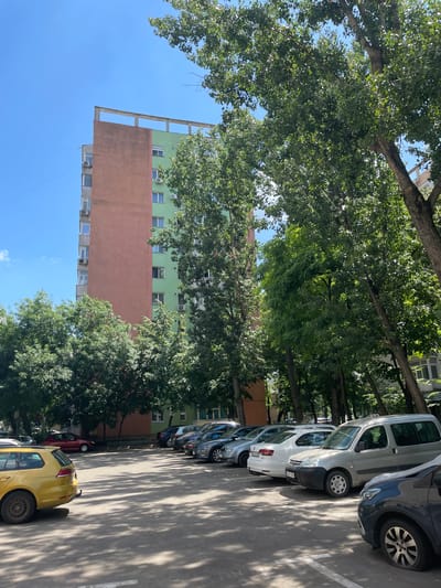 The image depicts a street scene in Iași, Romania, on a sunny day. A line of parked cars, including a yellow hatchback, a dark blue sedan, a silver van, and a white hatchback, are visible on an asphalt surface marked with white lines. Lush green trees with full foliage cast dappled shadows on the ground and the cars. In the background, a multi-story building with a pale pink lower section and a green upper section stands against a clear blue sky. The building features numerous windows and air conditioning units. The orientation of the image is landscape, but it is rotated 90 degrees counter-clockwise. No people are visible in the frame. The scene suggests a typical urban environment during daytime.