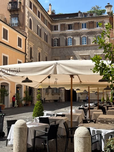 The image shows an outdoor dining area of a restaurant in Rome, Italy. The scene is dominated by a large, cream-colored awning that stretches across the frame, with wooden poles supporting it. Below the awning, tables are set with white tablecloths and glassware, suggesting a meal is about to be served or has recently concluded. Dark chairs are arranged around the tables.
In the background, traditional Roman architecture is visible, with stone buildings featuring arched windows and ornate facades. Green foliage from trees and plants adds a touch of nature to the urban setting. The time of day appears to be daytime, with sunlight casting shadows on the cobblestone street. The weather seems pleasant, as indicated by the outdoor dining setup.
One notable detail is the text visible on the awning: "pierluigi ROMA 1938". This clearly identifies the restaurant and its founding year. A few people are present, including a person in a red top walking away from the camera in the background. The overall atmosphere seems to be one of a quiet, picturesque Roman street.