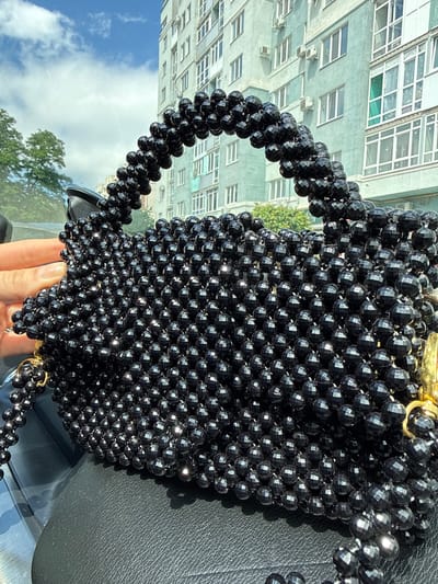A black beaded handbag rests on a black leather surface, possibly a car seat. The handbag is intricately covered in faceted black beads, giving it a textured and reflective appearance. A woman's hand, with manicured nails, is visible on the left side, gently holding the bag. In the background, a light-colored apartment building with multiple windows stands against a blue sky with scattered clouds. Trees are visible to the left of the building. The overall scene suggests an outdoor setting, possibly in a residential area within the city of Poltava, Ukraine, given the architectural style of the building and the context provided.