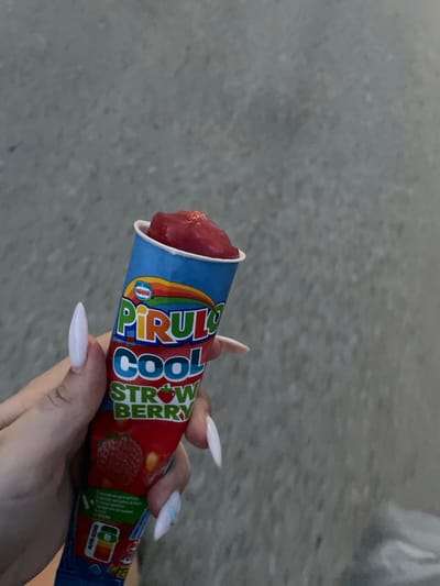 In Skala Avdiron, Greece, a hand with long, pointed, pale pink nails holds a bright blue and red packaged strawberry-flavored frozen dessert. The packaging features the "Nestlé" logo and the brand name "PIRULO COOL" in large, colorful letters, with "STRAWBERRY" written below, accompanied by an illustration of a strawberry. The frozen dessert itself is a vibrant red, protruding from the top of the packaging. The background consists of a textured, grayish surface, possibly a sidewalk or pavement, suggesting an outdoor urban or semi-urban setting. The lighting is natural, with no artificial light sources clearly visible.