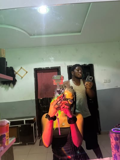 The image depicts an indoor scene in Ipetumodu, Nigeria, captured as a mirror reflection. The primary subject is a female, positioned centrally and seen from the torso upwards. She has dark skin, braided dark hair styled in multiple cornrows/braids, and wears clear-framed glasses. She is dressed in a dark, possibly strapless, garment. Her fingernails are painted red, and she wears black scrunchies on both wrists, along with a gold-colored necklace featuring a heart-shaped pendant. She holds a smartphone with a patterned case in both hands, angled to capture the mirror selfie. A pink/purple charm, resembling a spiked object, hangs from the phone. A strong, multicolored light refraction (red, orange, green, yellow) is cast across her upper body.
Behind her and to the right, a male individual is reflected. He has dark skin, short dark hair, and wears an earring in his left ear. He is dressed in a light-colored sleeveless tank top and dark shorts. He holds a dark-colored smartphone in his right hand, and his mouth is slightly open.
The background reveals a room with walls painted light green/mint green on the upper section and dark grey on the lower section, separated by a horizontal line. Numerous small, reflective speckles are visible on the upper wall. The white ceiling features a recessed panel and a single bright circular overhead light fixture. Two dark doorways are present; the left doorway is partially obscured by a dark red curtain or fabric, while the right doorway shows a white electrical switch plate on the adjacent wall.
Various objects are also reflected: On the left, a pink shelf holds a tall, gold cylindrical object and dark hair extensions. Below the shelf, a wooden, foldable drying rack is visible, alongside a dark cabinet with metallic accents and a translucent container with a printed label. On the right, a cylindrical object with a camouflage pattern (pink, black, grey) and glowing purple illumination, likely a portable speaker, rests on a surface.