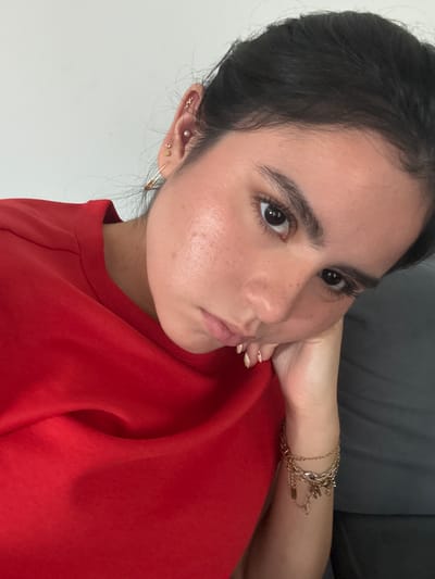 A selfie of a young woman with dark hair pulled back is angled diagonally. She wears a red shirt and multiple gold bracelets on her right wrist. Her right hand is positioned behind her head, supporting her chin. She has multiple earrings in her left ear. The background is split between a white wall and a gray surface. The photo appears to be taken indoors with natural lighting. There's no explicit information to determine that the photo was taken in Austin, United States of America.