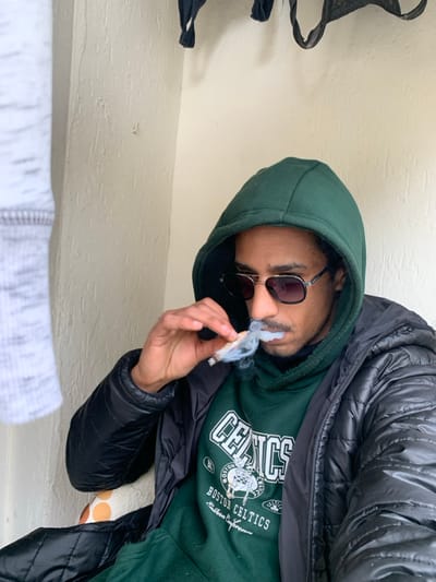 The image features a person, likely male, in a casual setting, shown from the chest up. The image is oriented vertically, with the person's head tilted to the right, as if leaning back or captured from an unusual angle. The person is wearing a dark green hooded sweatshirt, with the hood up, partially covering their head. Over the sweatshirt, they are wearing a black puffy jacket. They have dark sunglasses on, obscuring their eyes. A slight beard or stubble is visible on their chin and upper lip.
The central activity involves the person holding a lit hand-rolled cigarette or joint between the thumb and forefinger of their right hand, which is brought up towards their mouth. Wisps of smoke are actively emanating from the cigarette/joint and from around the person's mouth, suggesting they are in the process of smoking or exhaling.
On the front of the green sweatshirt, there is visible text and a logo. Prominently displayed is "CELTICS" in large white lettering. Below this, smaller text reads "Boston Celtics" and "Eastern Conference." A graphic resembling a basketball team logo, specifically the Boston Celtics' leprechaun mascot, is also present on the sweatshirt.
The background appears to be a plain, light-colored wall or surface, possibly cream or off-white, with some subtle texture. In the upper left corner of the image, part of a black fabric item, possibly a bag strap or clothing, is visible. In the bottom foreground, a blurry portion of a light-colored, possibly white and grey patterned fabric (like a towel or blanket) is present.
The overall impression is casual and relaxed, with the person engaged in smoking. The time of day and specific location are not discernible from the image.
