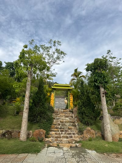 The image shows an oriental-themed gateway in a garden-like setting, located in the city of Liên Nghĩa, Vietnam. The gateway, constructed of tan-colored brick, is painted lime-green with yellow lettering. It is nestled between two tall trees, each with light gray bark. The gateway is bordered by green vegetation and rocks. A patterned concrete walkway lies adjacent to the gateway. The sky above is cloudy with pale blue patches.
