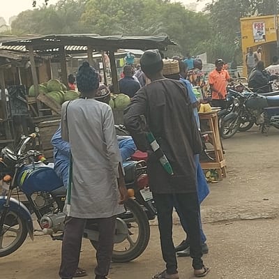 The image displays an outdoor street scene, bustling with people, motorcycles, and vendor stalls under natural daylight. The original image is rotated 90 degrees clockwise; this description will treat the image as if it were correctly oriented with the ground at the bottom.
In the foreground, two men are prominently featured, viewed from behind. The man closer to the right wears a light grey, long-sleeved tunic over dark trousers and sandals, topped with a dark, possibly patterned or braided, cap. To his immediate right, a distinct green and white striped object, resembling a stick or a rolled item, is visible. Directly in front of him, another man, also seen from the back, wears a dark brown or black long-sleeved tunic over dark trousers and sandals, with a light brown woven cap. He also has a similar green and white striped object tucked into his back.
Between these two men and extending to the left, a wooden vendor's stall or cart is present, laden with numerous green, round fruits that appear to be coconuts or small watermelons. Another person in blue attire is partially obscured behind this stall.
The mid-ground features several motorcycles, with a blue one visible towards the left and others scattered throughout. Various individuals are standing or walking around, engaged in typical street activities. One person, to the left of the prominent foreground figures, is visible in a bright orange long-sleeved shirt.
In the background, more people and motorcycles are discernible, indicating a busy thoroughfare. To the far upper left, a yellow structure, possibly a kiosk or a small shop, is visible. On this structure, the letters "POS" are clearly legible, likely indicating a Point of Sale service. The ground appears to be unpaved, possibly dirt or a rough, dusty surface. Sparse vegetation, like trees or bushes, can be seen in the distant background on the left side.
The overall atmosphere suggests a casual, everyday market or street environment, likely in a region where such attire and informal vending are common. The lighting indicates it is daytime, possibly overcast or hazy, contributing to the soft shadows.