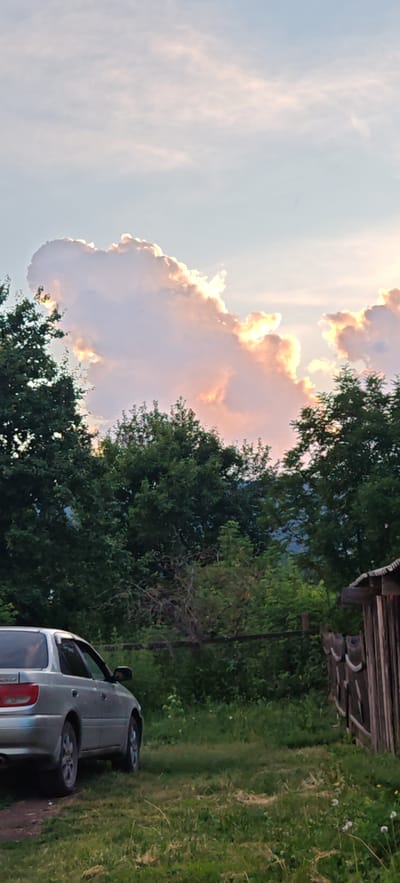 In the city of Altayskoye, Russia, a silver sedan is parked on a grassy area. The car is positioned to the left of the frame, facing forward. Behind the car, dense green trees form a natural backdrop. The sky above is a pale blue, with large, luminous clouds tinged with pink and orange hues, indicating either sunrise or sunset. To the right of the image, a wooden structure, possibly a shed or fence, is visible, constructed from weathered planks. The ground is covered in grass and some scattered wildflowers. The overall scene suggests a rural or semi-rural setting. There are no people visible in the image.