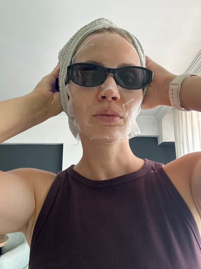 The image shows a person with a towel wrapped around their head, wearing sunglasses and a sheet mask. They are holding their hands to the sides of their head. The person is wearing a dark purple ribbed tank top. A light pink smartwatch is visible on their left wrist. The background appears to be an indoor setting with light-colored walls and windows covered by white curtains. There is a dark gray wall in the background on the left side of the image. There is no indication of the location in the city of Fethiye, Turkey.