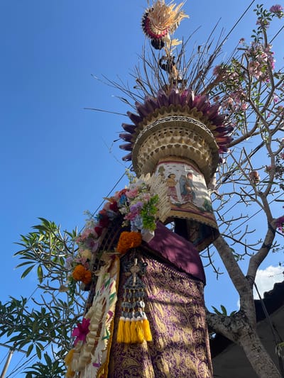 This low-angle, full-shot image shows an intricately decorated, multi-tiered structure resembling a totem pole, possibly part of a Balinese ceremony or offering. The structure is adorned with vibrant artificial flowers in shades of pink, orange, red, blue, and white, as well as gold tassels and woven decorative elements. A richly patterned purple and gold fabric wraps around the base of the structure, with intricate gold motifs. The top of the structure features layered, fan-like elements in shades of purple and cream, topped with a headdress-like decoration of woven straw or reeds. A painted scroll or banner depicting two figures in traditional attire wraps around a middle section. The structure is set against a clear, bright blue sky. To the right, the bare branches of a tree with small pink flowers are visible, and to the left, a tree with large green leaves is present. The overall impression is one of vibrant color and detailed craftsmanship, set against a natural outdoor environment. The location is identified as Ubud District, Indonesia.