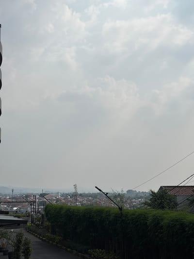 The image shows a cloudy sky over a residential area in Bogor, Indonesia. In the foreground, there is a dark, paved road curving to the left, bordered by a yellow line. To the right of the road is a dense, dark green hedge of bamboo plants. Further back, a building with a red tiled roof and grey walls is visible. Across a valley, a densely packed residential area with many red-tiled roofs stretches out, interspersed with trees. Several cell phone towers are scattered throughout the cityscape. The sky is predominantly light grey with some scattered white clouds, suggesting an overcast day. On the far left of the image, a portion of a tall, dark, curved structure, possibly part of a building or installation, is visible.