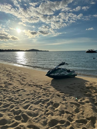 The image depicts a serene beach scene at Baan Mae Nam, Thailand, likely in the late afternoon given the warm, angled sunlight casting long shadows across the sand. The sky is a mix of blue and white, with scattered, wispy clouds that are illuminated by the sun, creating a sense of a pleasant, clear day.
The foreground is dominated by a sandy beach, marked with numerous footprints, indicating recent activity. A dark-colored jet ski, with green accents and some visible white text that appears to read "jet ski" and other brand-specific markings, rests on the sand near the water's edge.
The middle ground features the gentle lapping of waves on the shore, with the sun's reflection shimmering on the water's surface, creating a bright, almost dazzling effect. Further out in the sea, a few small boats or buoys are visible, hinting at marine activity or leisure. In the distance, a hazy, mountainous silhouette suggests the island's topography.
The overall mood conveyed is one of tranquility and relaxation. The lighting and natural elements suggest a peaceful moment on a tropical beach, ideal for water sports or simply enjoying the coastal ambiance. There are no people visible in the frame.