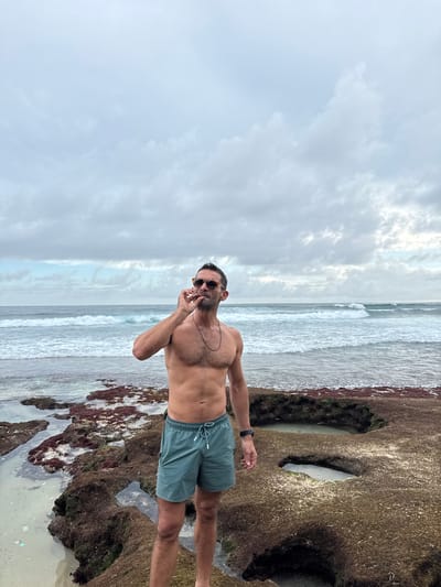 The image depicts a fair-skinned, shirtless man wearing dark sunglasses and green swim trunks standing on a rocky beach at Pecatu, Indonesia. He has a thin necklace and a watch on his left wrist, and appears to be holding something small between his fingers near his mouth. Behind him is the ocean with waves breaking onto the shore. The sky is overcast with dense clouds. The rocks in the foreground are covered with what appears to be reddish-brown algae.