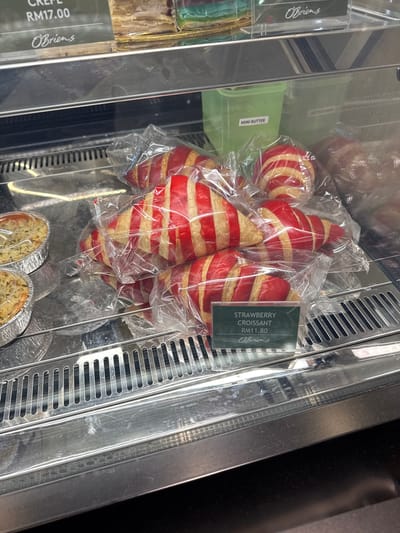 The image shows a close-up of a refrigerated display case in a food establishment, likely in Kuala Lumpur, Malaysia, given the currency "RM" which stands for Malaysian Ringgit and the brand "O'Briens" which is a known cafe chain in Malaysia. The display case contains various baked goods. In the foreground, there are several individually packaged croissants with red and yellow stripes, suggesting a strawberry flavor. A small green sign in front of these croissants reads "STRAWBERRY CROISSANT RM11.80 O'Briens". To the left, there are two foil-lined containers of what appear to be baked goods, possibly quiches or small pies, also packaged. In the background, there are some green plastic containers, one labeled "MINI BUTTER". The overall environment appears to be a commercial food retail setting, with polished stainless steel surfaces and the reflective glass of the display case. The lighting is bright, illuminating the products. No people are visible in the image.