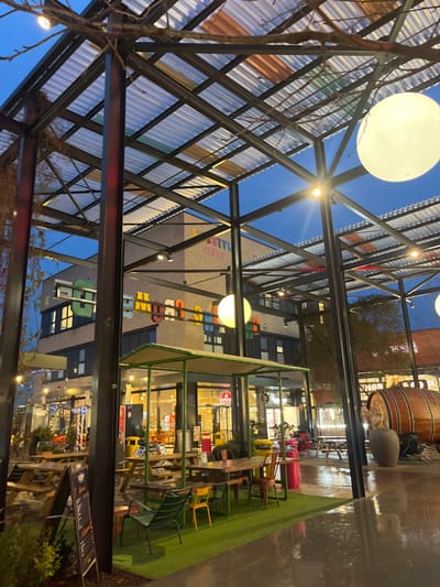 The image displays an outdoor commercial and leisure space at dusk or early evening in Reichstett, France, identified as a "Lifestyle Center" by prominent signage. A large, open-air metal truss structure with a corrugated translucent roof, partially incorporating colored panels (orange, green, clear), spans the foreground and midground. Bare tree branches are intertwined within this overhead structure, which also supports multiple spherical white pendant lights and smaller, direct illumination sources.

In the background, a multi-story building constructed of light-colored brick features the text "LIFESTYLE CENTER" on an upper section and "MEUGANG" in large, colorful block letters centrally. Numerous windows on this building are illuminated from within.

At ground level, an outdoor seating arrangement is present. To the left, several wooden picnic tables with benches are visible, with one individual faintly discernible in the distance near these tables. An A-frame chalkboard sign stands in the left foreground. A central section of the ground is covered with green artificial turf, hosting various tables and chairs in bright colors including green, yellow, orange, and red. This turf area is partially sheltered by a smaller, green-canopied structure. Beyond this, through glass storefronts, interior restaurant spaces are visible, including a red sign that partially reads "BURGER." Red cylindrical trash bins are located near the seating.

On the far right, a large wooden barrel rests horizontally on the wet, paved ground. Adjacent to it stands a sculptural figure, resembling a rooster, positioned atop a dark pedestal. The ground throughout the visible area appears wet, reflecting the artificial lights. No direct interactions between individuals are clearly depicted beyond the single discernible person in the distance.