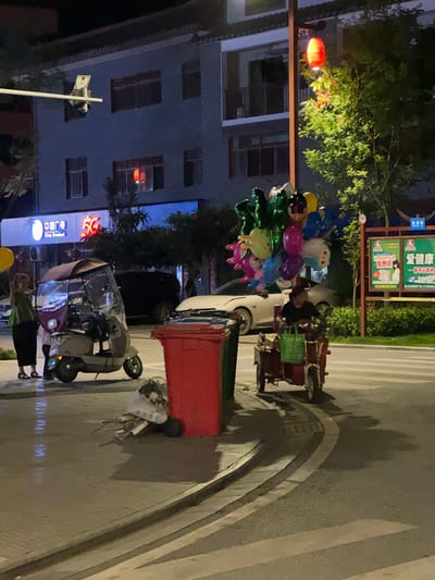 In Eshan Yi Autonomous County, China, at night, a street scene unfolds. A woman in a green top and black skirt stands beside a three-wheeled scooter with an umbrella canopy. The scooter is light beige and has a dark purple cover over the passenger seat. Further to the right, a person in a dark shirt rides a red three-wheeled vehicle, carrying a bunch of colorful balloons that reach high above their head, including green, pink, blue, and yellow balloons, some with character designs. A large red trash bin is positioned to the left of the red vehicle. In the background, a building with multiple windows is illuminated, and a red lantern hangs from a pole. A street sign with Chinese characters is visible on the right. The road surface shows a pedestrian crossing.