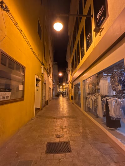 The image depicts a narrow street in Blanes, Spain, illuminated by streetlights at night. The street is paved with cobblestones and flanked by buildings. On the left, a building with a vibrant yellow facade features a large window with a "For Rent" sign. Further down the street, a building with a dark facade displays several signs for "OS Serveis Immobiliaris." On the right, a shop with a large display window showcases an array of clothing items. The street is empty except for two figures walking in the distance and bicycles parked outside a building.