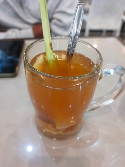 A glass mug filled with brown liquid sits on a light-colored surface. A green stalk protrudes from the drink along with a metal spoon. The drink seems to contain pieces of something at the bottom. In the background, a person's upper body is blurred along with a phone and a white chair. The scene suggests a cafe setting with someone enjoying a beverage.