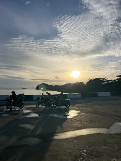 The image captures a serene coastal scene at sunset, possibly in Thailand given the implied context. The foreground shows a wet asphalt surface with several puddles reflecting the warm light of the setting sun. Three individuals on motorcycles are visible, silhouetted against the bright sky. One motorcycle on the left has a green delivery box attached to its rear. The middle motorcycle carries two people, and the third motorcycle on the right appears to have one rider. Behind the motorcycles is a concrete barrier with alternating black and white sections, separating the road from the body of water. The water stretches out to the horizon, where a large vessel, likely a ferry, is visible in the distance. Lush green trees and a hilly island form the backdrop on the right side of the image, with the sun setting behind them, casting a warm glow and creating lens flare. The sky is adorned with a dramatic display of clouds, ranging from wispy cirrus formations to more textured altocumulus clouds, all illuminated by the golden hues of the sunset. The overall atmosphere is tranquil and picturesque.
