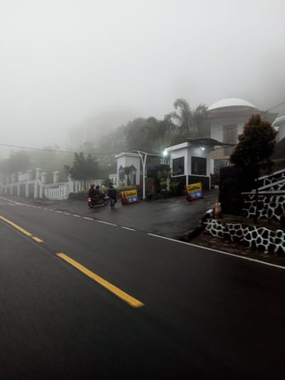 The image depicts a foggy roadside scene in Dawathang_Dorjibi_ Kashingtsawa, Bhutan, under diffused lighting conditions. A wet, dark asphalt road, marked with a dashed yellow center line and a solid white right-edge line, occupies the foreground. On the left, a white ornamental fence lines the road, behind which is dense green foliage and a white structure featuring an arched entrance. Further back and partially obscured by fog, another white building and a sign displaying "ATTA" are visible on a hillside. In the midground, two individuals are present: one person is seated on a dark motorcycle, wearing a red helmet, facing towards the right. Another person, dressed in dark clothing, walks alongside the motorcycle, also facing right. Adjacent to the road on the right, a low wall constructed of dark stones with white mortar patterns is visible, alongside an orange traffic cone. Two yellow and blue temporary signs stand near the road. Behind these elements, a white building with a distinct domed roof is partially visible amidst trees, along with a smaller white booth-like structure. The background consists of heavily fog-obscured terrain.