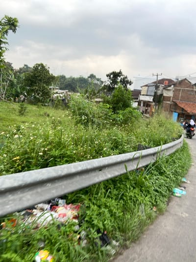 The image shows a view of a road in the city of Kecamatan Margaasih, Indonesia, located at latitude -6.963 and longitude 107.545. The road is paved and curves to the right. A metal guardrail is placed along the edge of the road, separating it from a grassy area with overgrown vegetation. The guardrail is partly covered by litter, including plastic bags, bottles, and other debris. There is a single motorcycle parked on the road to the right, out of focus. There is a building structure in the background, out of focus, suggesting an urban setting. The sky is overcast, and the vegetation is green.