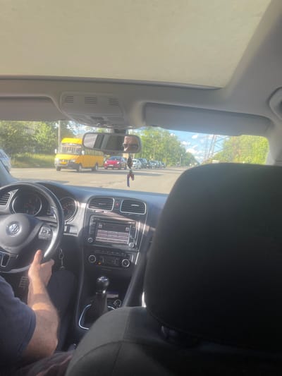 The image shows the interior of a Volkswagen car, viewed from the passenger seat. The driver's hands are on the steering wheel, and their arm is visible. The car's dashboard, including the radio and gear shift, is clearly visible. Outside the car, there is a street scene with other vehicles, including a yellow bus and a red car. Trees line the street, and the sky is blue with some clouds. The scene appears to be in Poltava, Ukraine, given the presence of a yellow bus with signage that suggests it is a route taxi.