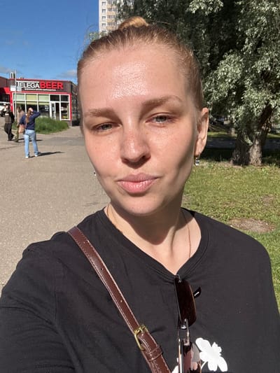 A woman with blonde hair pulled into a bun takes a selfie outdoors on a sunny day. She is wearing a black t-shirt and a brown crossbody bag. Her face is turned towards the camera, and she has freckles and light makeup. Behind her, a building with red and white signage that reads "ELEGA BEER" is visible, along with a few people walking on a paved area. A grassy area with trees is also present in the background. The image appears to have been taken in Rusanovo, Russia.
