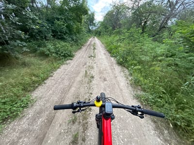 The image is a first-person perspective shot from a bicycle on a dirt road. The road stretches into the distance, flanked by dense green vegetation on both sides. The bicycle's handlebars, a red frame, and a cycling computer mounted in the center are visible in the foreground. A small yellow rubber duck is attached to the handlebars. The road surface is uneven, with visible tracks and patches of grass. The sky is partly cloudy. The location is Vetren, Bulgaria based on prior user data.