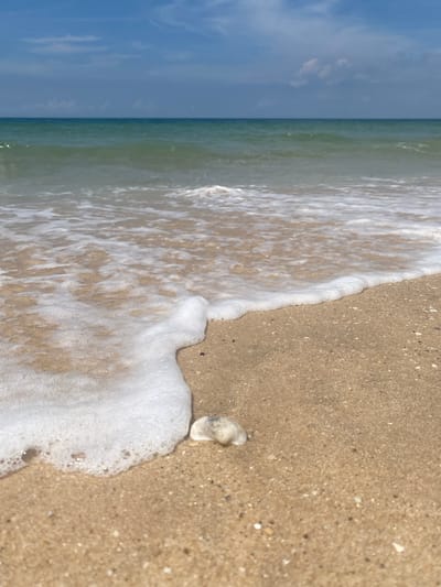 The image depicts a close-up, low-angle view of a sandy beach in Phu Thuy Ward, Vietnam. The foreground is dominated by wet sand, with a gentle wave of white foam from the ocean washing ashore. A small, light-colored object, possibly a shell or piece of coral, rests on the sand near the edge of the foam. The middle ground features the tranquil, turquoise ocean with subtle ripples and a clear horizon line. The background shows a bright blue sky with scattered white clouds. The scene conveys a peaceful and serene coastal environment.