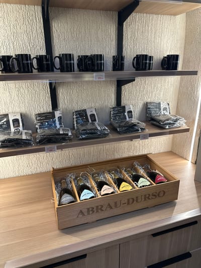 The image displays a retail setting, likely a shop or boutique, within Abrau-Dyurso, Russia. The scene is divided into two main levels, both featuring merchandise presented on wooden shelves. The upper shelf holds several black mugs, each bearing a white inscription, possibly a brand logo. To the right of the mugs, a price tag displays "950 руб." The middle shelf contains black, individually packaged items, likely clothing or accessories, in transparent plastic wrapping. Each package has labels with text and some feature the letter "M". A price tag on this shelf reads "1900 руб." The bottom level showcases a wooden crate filled with bottles of sparkling wine. The crate is branded with the embossed text "ABRAU-DURSO". The bottles are arranged in a row, nestled in shredded white paper. The overall environment is clean and well-lit, with a textured wall in the background. There are no people visible in the image.