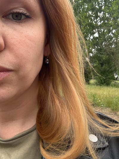 The image shows a close-up, cropped view of a person's face, focusing on their left eye, cheek, and mouth. They have fair skin with freckles and green eyes with visible eyelashes. A small, light blue earring with a rectangular stone is visible on their left ear. Their hair is a prominent feature, with strands of light brown or auburn hair cascading across the frame. The background is blurred, suggesting an outdoor environment with green trees and vegetation, possibly a park or natural area. The person is wearing a green t-shirt and a black leather jacket. The scene does not provide any specific clues about the city of Заволжье, Russia.