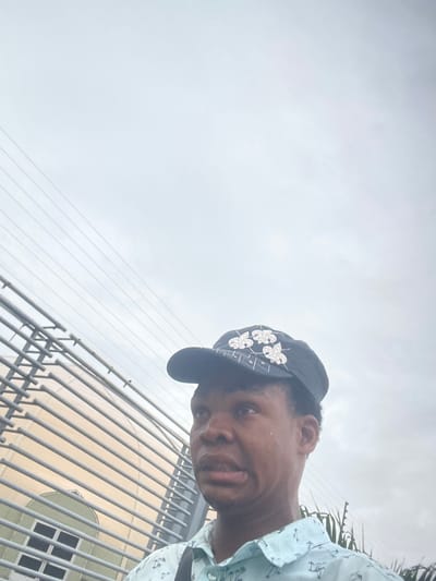 In Igboefon, Nigeria, a close-up shot captures a young Black person wearing a black baseball cap adorned with white floral appliques. The person is dressed in a light blue collared shirt featuring a repeating pattern of palm trees and anchors. They are looking off to the left of the frame with a slightly distressed expression. In the background, horizontal metallic bars form a fence or railing, behind which a building with a light-colored facade and a section of green-tinted windows is visible. Overhead, a network of power lines stretches across an overcast sky. The composition is tight, focusing on the individual's face and upper torso, with the urban background providing context.