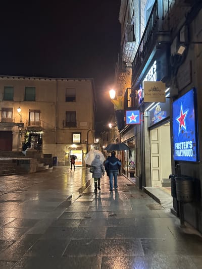 The image shows a wet, cobblestone street in Segovia, Spain, at night. Two people with umbrellas are walking toward a building. The building has a sign that reads "Foster's Hollywood" and a red star. The building is brightly lit. There are other people walking in the distance. There are two buildings in the background. The scene is lit by streetlights and the reflections from the buildings. The street is wet from recent rain. The image was taken near the Plaza Mayor, Segovia, Spain (40.949, -4.122).