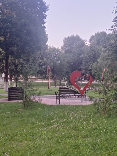The image depicts a park scene in Kapyĺ, Belarus. In the foreground, a green lawn is dotted with small white flowers. A paved walkway leads to a bench with an ornate black metal frame, featuring a large red heart-shaped sculpture as the backrest. To the left of the bench, a small evergreen tree stands on the grass. Further back, a wooden trash receptacle is visible next to the walkway. The background is dominated by mature trees with dense green foliage under a hazy, light-colored sky. The overall impression is of a quiet, public outdoor space.