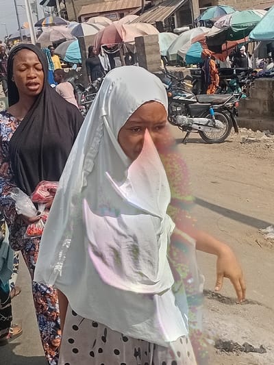 Two women pose in a bustling outdoor market. One woman, wearing a black hijab, looks directly at the camera with a slightly open mouth. Another woman, partially visible, is draped in a white hijab. A hand reaches out in the background toward a weathered wall. Motorbikes, stalls, and other shoppers create a chaotic market scene in the background. Umbrellas provide shade to the market stalls. The scene suggests a lively marketplace atmosphere.