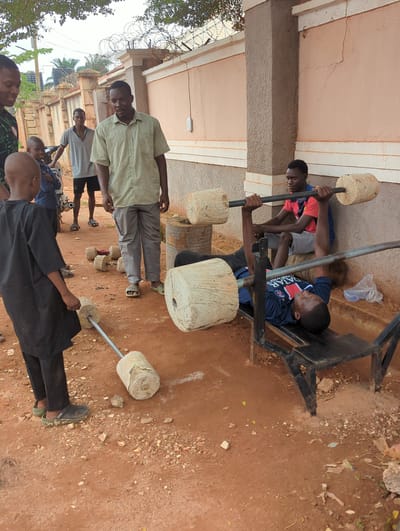 This image captures an outdoor scene in Unguwar Rimi, Nigeria, depicting a group of young men and boys engaged in physical activities and socializing.
In the foreground and midground, two young men are actively lifting weights. One young man, wearing a dark blue sports shirt with "QATAR AIRWAYS" and a PSG (Paris Saint-Germain) logo, is lying on a makeshift metal weight bench, pressing a barbell. The barbell is constructed from a metal rod with cylindrical concrete or stone weights at each end. Another young man stands nearby, holding a similar homemade barbell, ready to lift.
Several other individuals are present in the scene. A man in a light green plaid shirt and dark trousers stands observing, looking towards the camera. Further back, another person in a light grey shirt stands near a small motorcycle. Other figures are partially visible in the background, including boys.
The setting is an open area with reddish-brown dirt ground, scattered with small rocks and debris. The exercise takes place in front of a building with light-colored walls and a darker, textured lower section. Barbed wire is visible along a fence or wall in the background, suggesting an enclosed area. Some sparse greenery and trees are also visible.
The scene takes place during the day, under natural, diffused lighting. The notable detail is the improvised nature of the gym equipment, highlighting resourcefulness in creating fitness tools from available materials. The atmosphere is casual, portraying a communal fitness and social gathering.