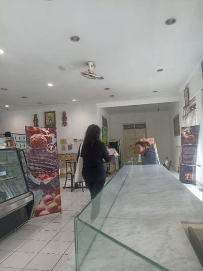 The scene is located inside a bakery shop in Purwokerto, Indonesia, at coordinates -7.428 latitude and 109.247 longitude. A woman in a black shirt and black pants is walking past a glass counter. She has a white bag on her left shoulder. Another person, possibly a man wearing a blue shirt, is seated at the counter. There is a large glass display case with an assortment of pastries inside. There is also a large poster advertising the pastries. The walls are white and the floor is tiled. There are ceiling lights and a ceiling fan in the room.