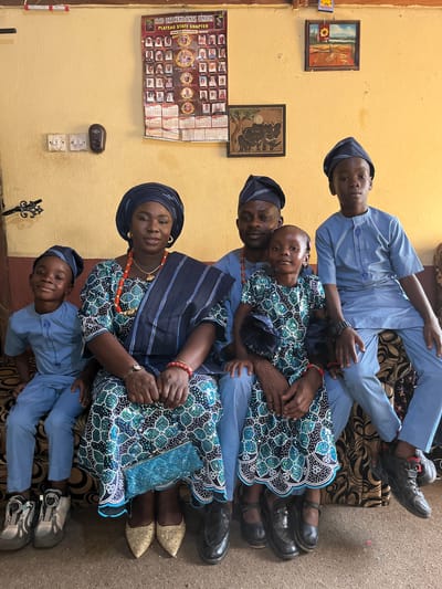The image shows a family portrait taken in a room with yellow walls. A woman in an ornate blue and green dress and headwrap sits centrally, adorned with a red beaded necklace and matching bracelets. To her right sits a girl in a matching dress and a man in a blue outfit and hat. Two boys wearing blue outfits are positioned above and below. The room features framed artwork, light switches, and a calendar on the wall. The location is Rafin Bauna, Nigeria.