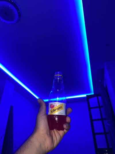 The image depicts an indoor scene in Baraki, Algeria, heavily illuminated by blue light. A right human hand, exhibiting moderate arm hair, is centrally positioned, holding a clear plastic bottle of Schweppes Premium Sparkling Grenadine containing a dark red liquid. The bottle's label, featuring pomegranates, is partially visible. The ceiling features a circular recessed light fixture with a metallic bezel in the upper-left corner, emitting blue light. Blue LED strip lighting traces the perimeter of the ceiling, casting a strong blue hue throughout the room. The walls reflect this blue illumination. On the right side of the frame, a dark, multi-paned rectangular structure, resembling a window or door frame, is visible. An obscure dark triangular form is present on the lower-left wall.