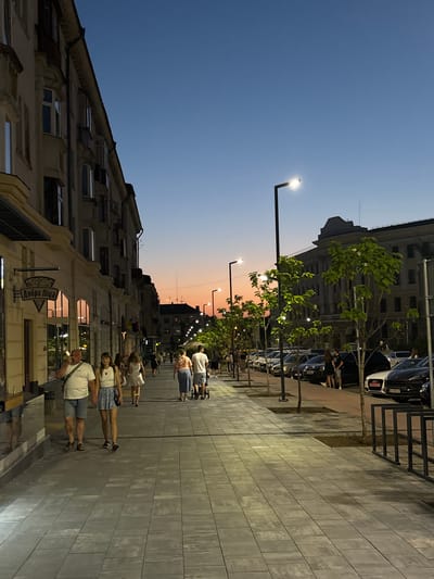 This media file depicts a street scene in Ternopil, Ukraine, during twilight. The image is oriented vertically, with the sky and buildings on the left and the sidewalk and people on the right. Several individuals are walking along the paved sidewalk, some in casual attire. Lampposts illuminate the walkway, casting a warm glow on the scene.
The sky transitions from a deep blue at the top to a soft orange and pink hue near the horizon, indicating the setting sun. Buildings line the street, with architectural details suggesting an urban environment. Trees with green foliage are interspersed along the sidewalk, adding a touch of nature to the cityscape.
A sign with Cyrillic text, "Добра піца," which translates to "Good pizza," is visible on one of the buildings, hinting at a commercial establishment. The overall atmosphere appears calm and relaxed, typical of an evening stroll. The weather seems pleasant, with no signs of rain or harsh conditions. The scene captures a moment of everyday life in Ternopil as day turns into night.