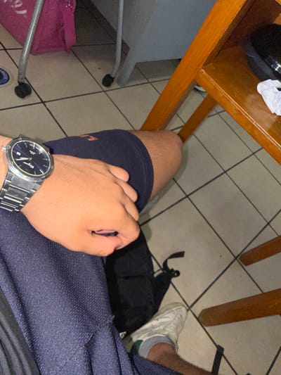 A first-person perspective image shows a person sitting with a silver wristwatch on their left wrist, resting on their thigh covered in navy blue fabric. Part of their hand is visible, as is a portion of their leg covered in a navy blue short. A white sneaker with green accents is visible on the right foot. A black backpack is situated near the leg. A wooden table with a white object and black object on it is in the upper left. The floor is tiled with light colored square tiles. The location is unknown and is not identifiable as Acapulco, Mexico based on the information available in the picture. A pink bag sits to the lower left.