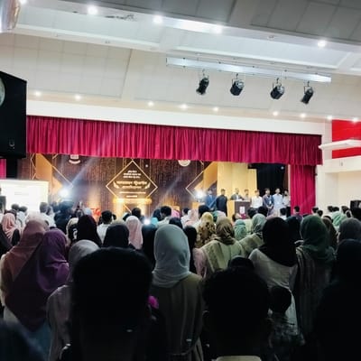 A large crowd is gathered in an auditorium for an event. On the stage, a group of people stands behind a podium. A backdrop displays the words "লালমোহন স্টুডেন্টস এসোসিয়েশন" in Bengali. The location is likely Lalmohan, Bangladesh, as the backdrop references the city. Most audience members are wearing modest clothing, and many women are wearing headscarves. Overhead lighting fixtures are visible. The crowd is the main focus of the image.