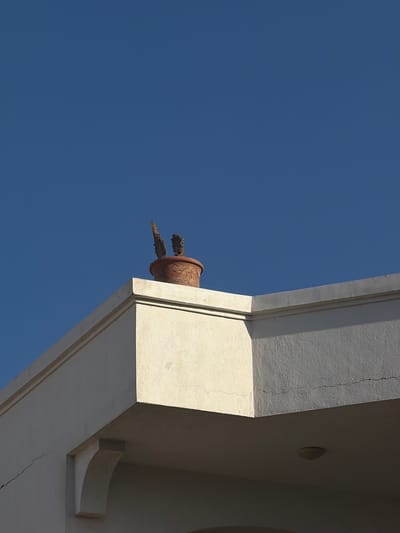 The image depicts a terracotta pot with two dried branches inside, placed on the edge of a building's roof against a clear blue sky. The building appears to be a white, stucco structure with architectural detailing, including an overhang and a decorative bracket. The pot has an intricate, embossed pattern around its rim. The scene is well-lit by sunlight, casting distinct shadows. The overall impression is one of stillness and a minimalist aesthetic, potentially located in a city like Sharm El Sheikh, Egypt, where such architectural styles and clear skies are common. There are no people or animals visible in the frame.