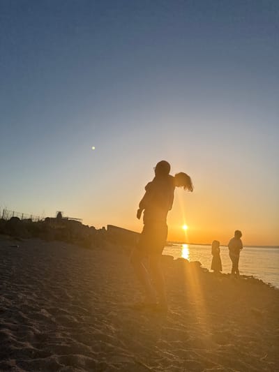 A silhouetted family is enjoying the sunset at a beach in Sozopol, Bulgaria. The scene is illuminated by the warm glow of the setting sun, which is low on the horizon and casting long shadows. A man is holding a child on his shoulders, while two other children stand nearby. The water is calm, reflecting the golden hues of the sky. The beach is covered in sand, with some rocks and vegetation visible along the shoreline. The overall mood is peaceful and joyous, capturing a moment of family connection during a beautiful sunset.