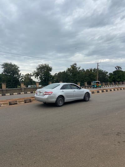 In Bar Jirgi Summa, Nigeria, a silver sedan is seen driving on a paved road under a cloudy sky. The car is positioned on the right side of the frame, facing away from the camera and moving to the left. To the left of the road, a sidewalk with black and white striped borders is visible, leading to a fence and a line of trees. In the background, beyond the trees, a small, colorful auto-rickshaw is also traveling on the road. Power lines are strung across the sky, and a single black object, possibly a bird or kite, is visible against the clouds.