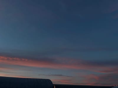 The image depicts a twilight sky over what appear to be industrial or agricultural structures in Novopokrovskaya, Russia. The sky transitions from a deep indigo-blue in the upper frame to lighter blue-gray hues, with distinct horizontal bands of stratiform clouds illuminated in soft pink and orange, consistent with a sunset. In the lower portion of the frame, two dark building silhouettes are visible. On the left, a large, curved, corrugated metal structure, possibly a quonset hut or hangar, occupies the lower-left. To its right, a lower, flatter roofline of another building extends along the horizon. Several small, faint, reddish-pink dots are present in the upper and mid-sky, consistent with lens flares or sensor artifacts. No individuals or discernible activities are present.