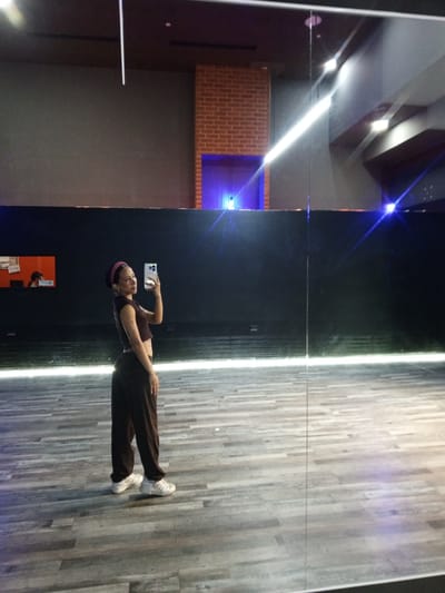 A full-body, medium-shot, selfie-style photograph taken in what appears to be a dance studio. The subject is a young woman standing on a wooden floor in front of a large mirror. She is wearing a dark crop top, dark loose-fitting pants, and white sneakers. A pink headband is on her head. She is holding a smartphone in her right hand, taking the picture. The background shows a dark wall with some lighting effects, a brick column, and reflections in the mirror. In the background, a small window reveals a person sitting at a desk. The lighting is dim, with bright streaks of light reflected from the overhead fixtures. The overall impression is a candid shot taken during a practice or rehearsal session. The location of Pampatar, Venezuela, is not explicitly visible in the image but has been provided as context.