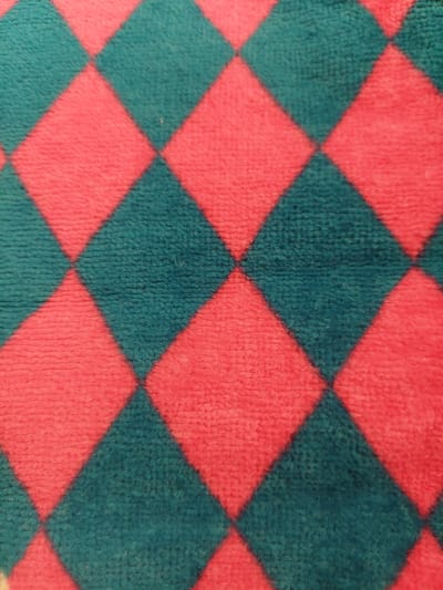 This is a close-up, overhead shot of a fuzzy textile with a diamond pattern. The diamonds are alternating between a bright pink and a teal color. The texture of the fabric is visibly soft and plush, suggesting it could be a blanket, rug, or similar soft furnishing. The lighting is even, and there are no discernible shadows, indicating it is likely shot indoors under artificial light or in an area with diffused natural light. The focus is sharp, allowing the texture of the fibers to be clearly seen. No people, objects, or specific setting are visible beyond the fabric itself. The image is abstract in nature, focusing solely on the repeating geometric pattern and the tactile quality of the material. There is no visible text in the image.